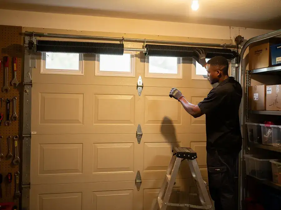 A technician on a ladder adjusts the torsion spring above a beige garage door.